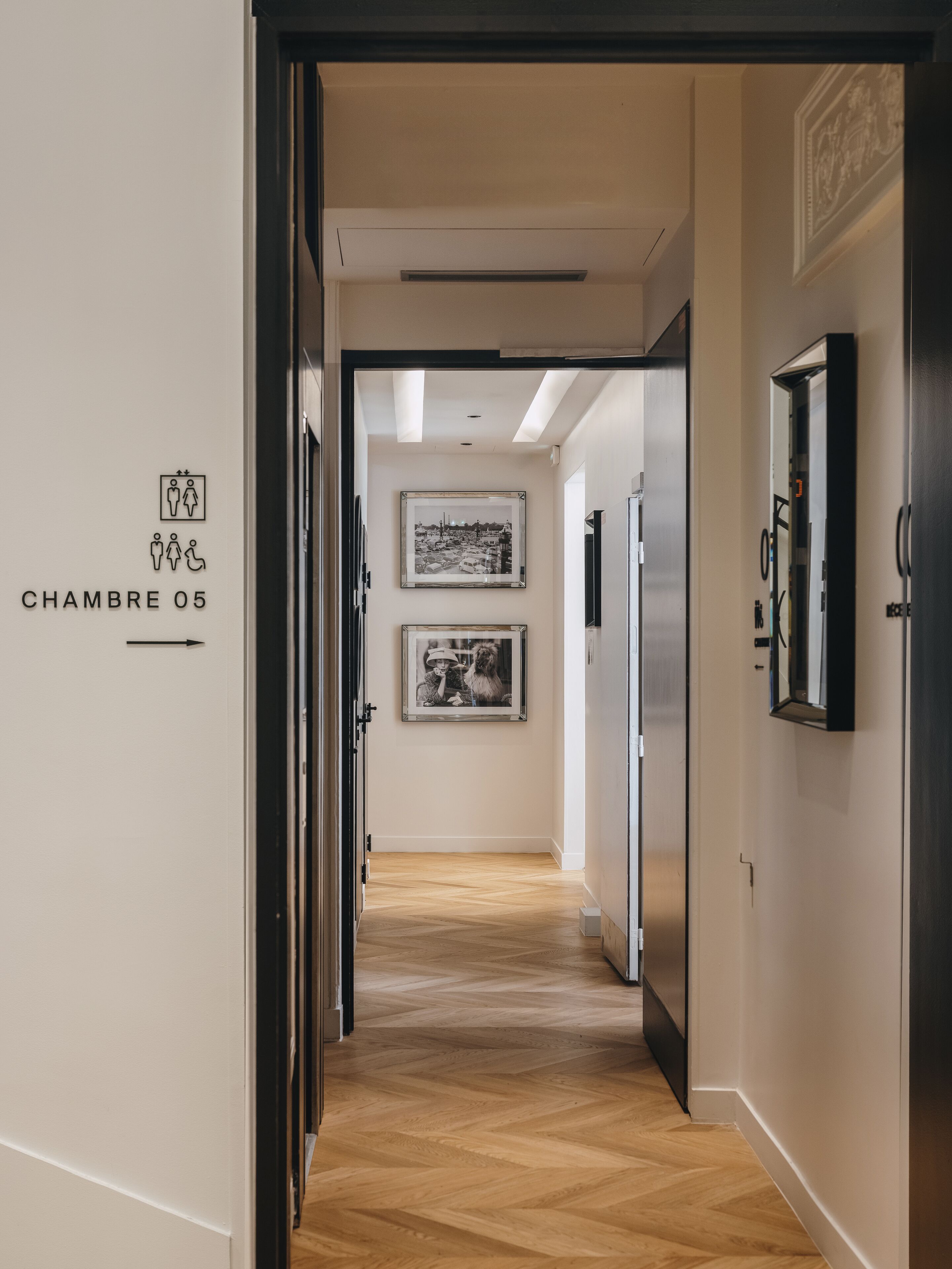 A hotel corridor with a wooden floor, two framed pictures on the wall, and a sign indicating 'CHAMBRE 05'.
