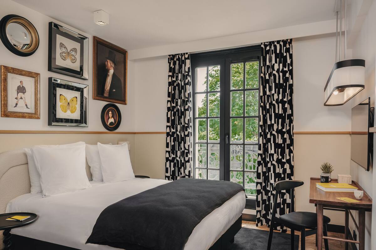 Une chambre d’hôtel avec un grand lit, un bureau avec une chaise, une fenêtre avec des rideaux à motifs et des tableaux encadrés accrochés au mur.