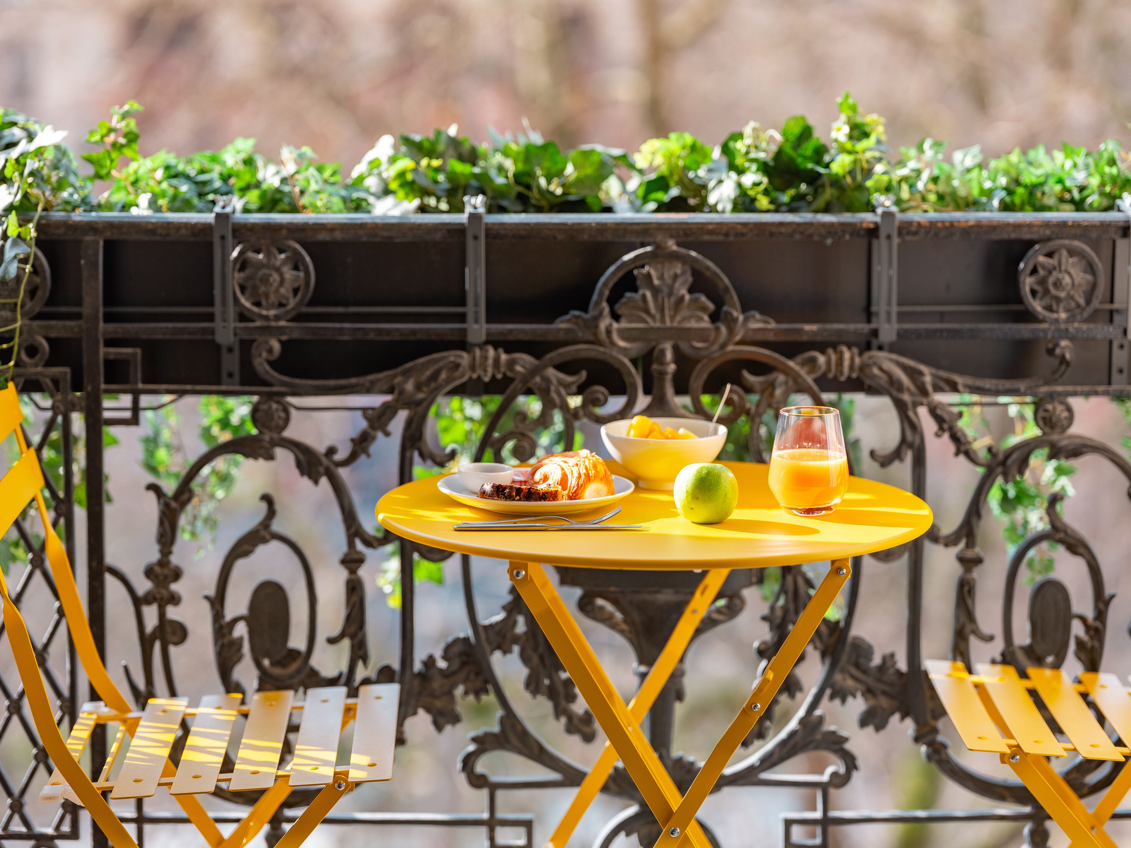 Une table jaune sur laquelle se trouve une assiette de nourriture, un verre de jus d’orange et un fruit vert. La scène se déroule sur un balcon orné de balustrades en métal et dont l’arrière-plan est composé de verdure.