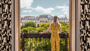 Una mujer de pie en un balcón con vista a un edificio histórico y abundante vegetación.