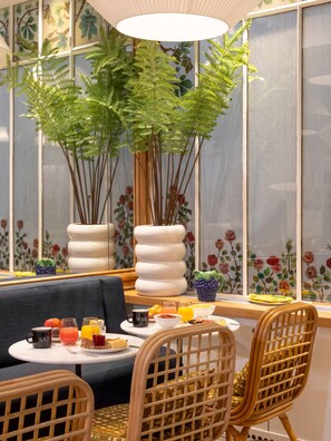 Un comedor con una mesa preparada para el desayuno, que incluye frutas, bebidas y una taza de café. Hay sillas de mimbre y un sofá.