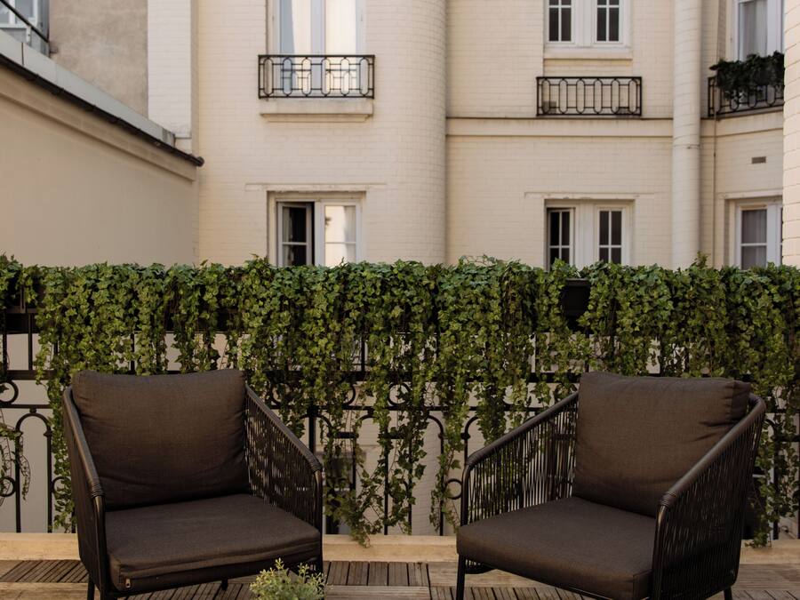 A terrace with two dark brown chairs and a wooden table, set against a building with multiple windows and balconies.