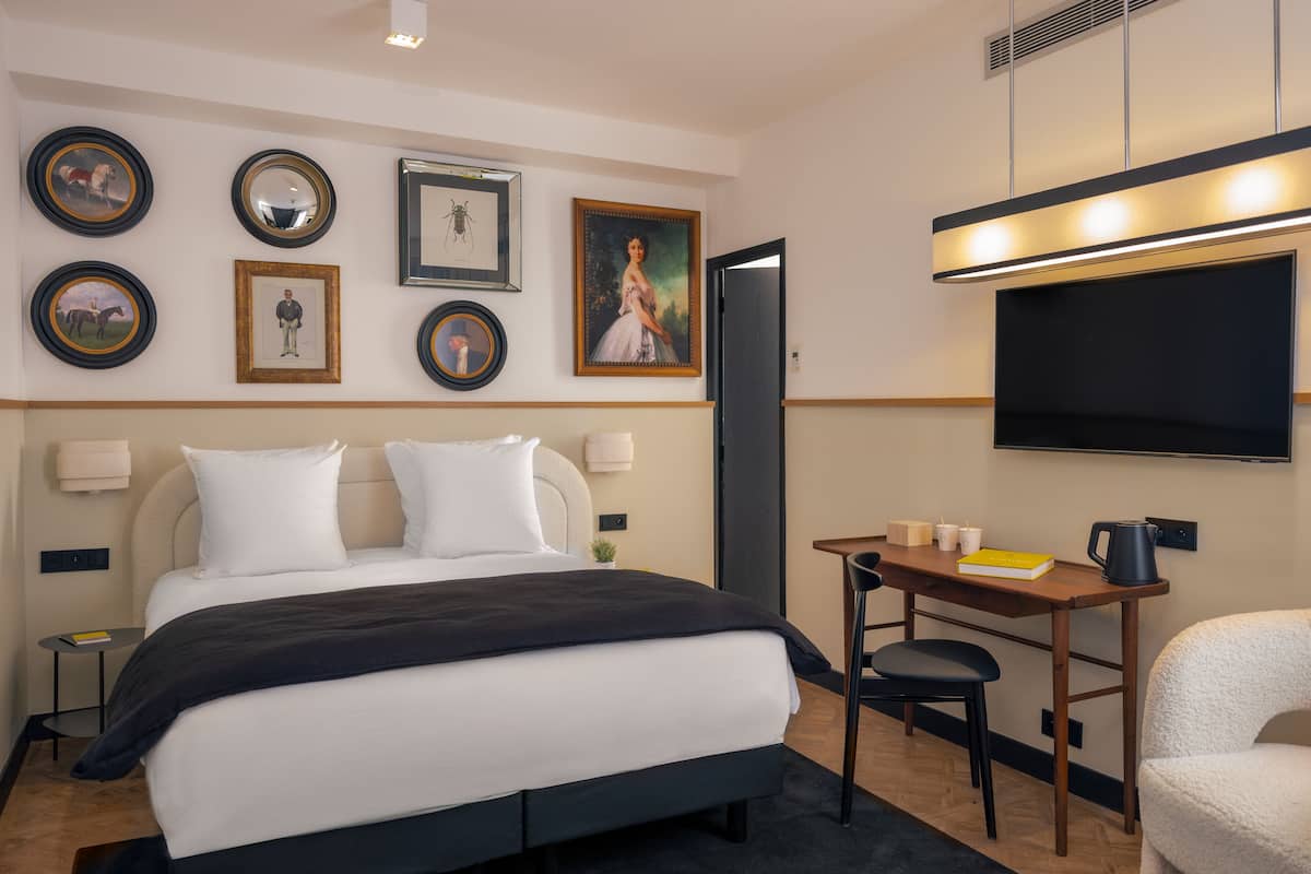 Une chambre d’hôtel comprenant un lit, des tables de chevet, un bureau avec une chaise, une télévision et des tableaux encadrés accrochés au mur.