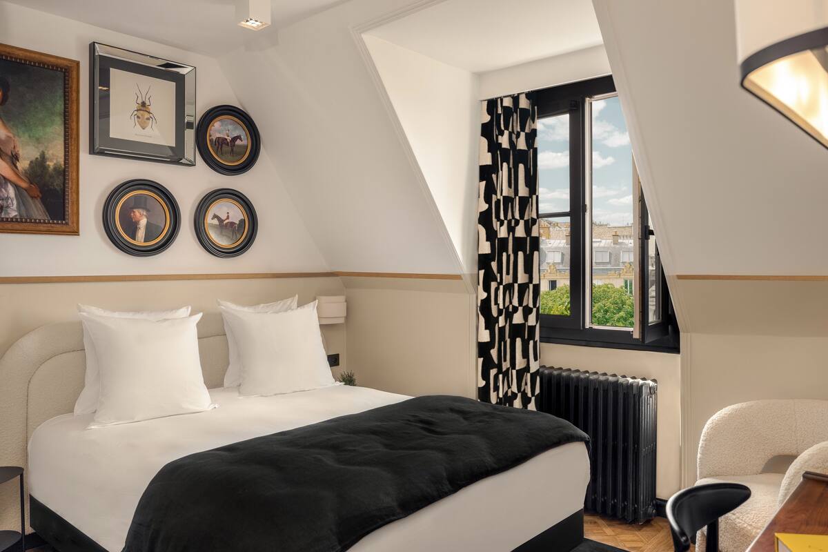Une chambre avec un grand lit, un radiateur, un bureau et une chaise. Les murs sont ornés de tableaux encadrés, et une fenêtre offre une vue sur les bâtiments environnants.