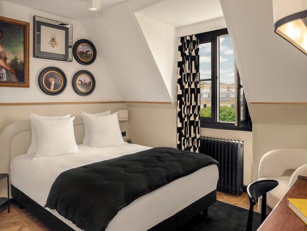 Un dormitorio con una cama grande, un radiador, un escritorio y una silla. Hay cuadros enmarcados en la pared y una ventana con vistas a edificios.
