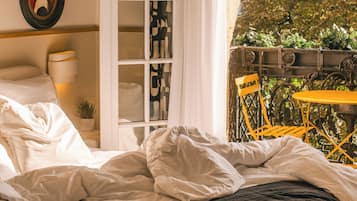 A bedroom with a bed, a wardrobe, a window with a view of a tree and buildings, and a balcony with a table and chairs.