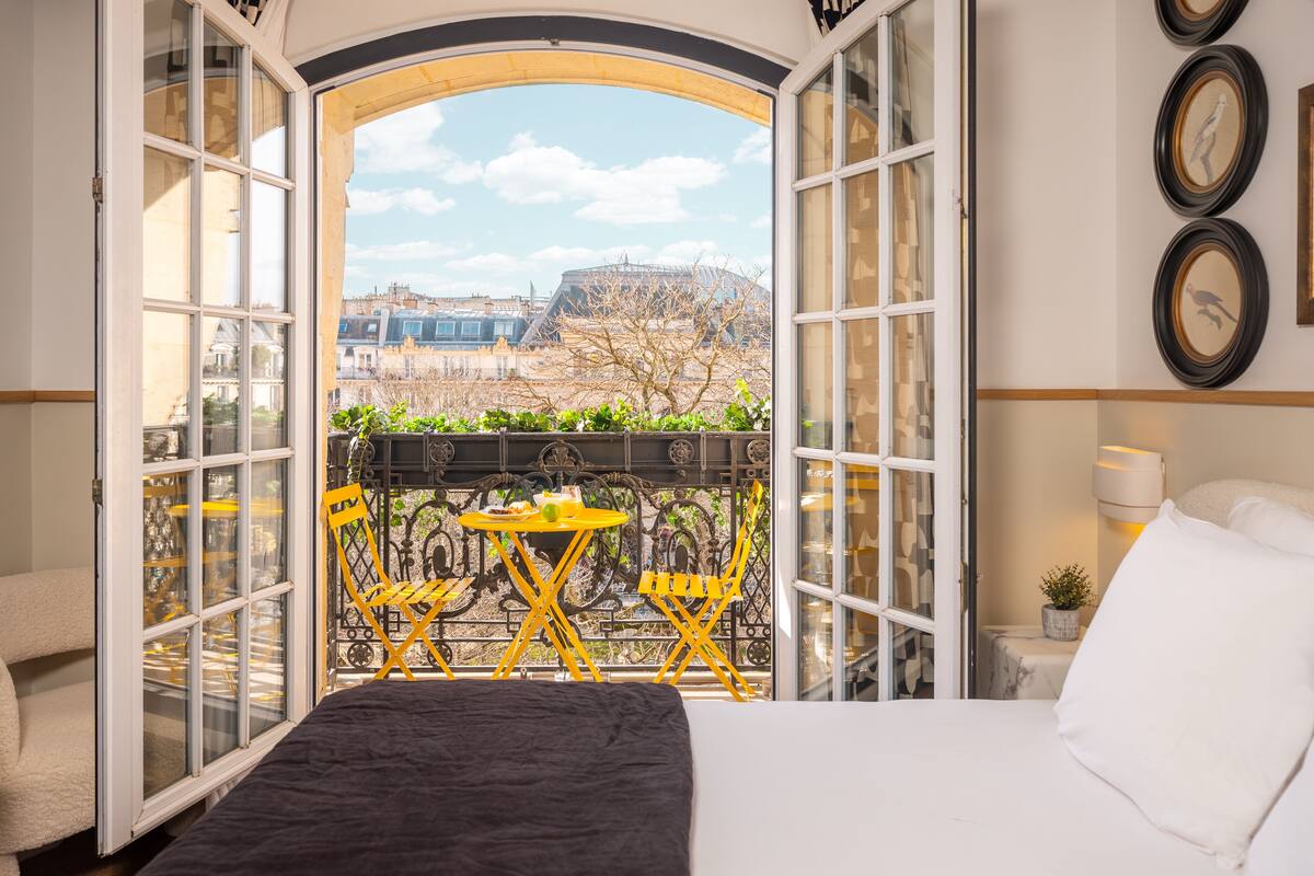 Une chambre d’hôtel avec un lit, un balcon doté de chaises jaunes et une vue sur des immeubles et des arbres.