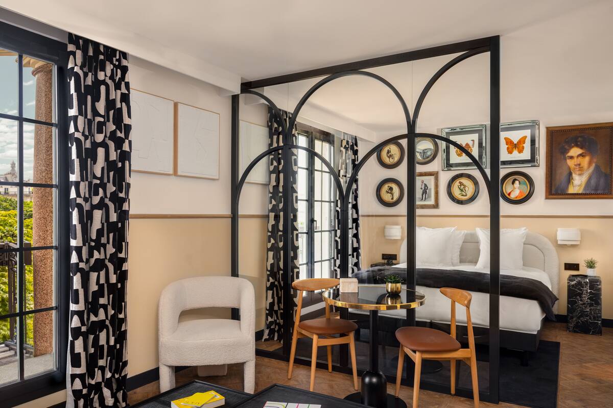 Une chambre d’hôtel moderne, équipée d’un lit, de deux fauteuils, d’une petite table et d’une grande fenêtre agrémentée de rideaux à motifs.