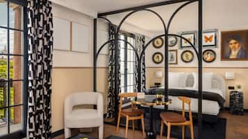 Una habitación de hotel moderna con cama, dos sillas, una mesita pequeña y un ventanal con cortinas estampadas.