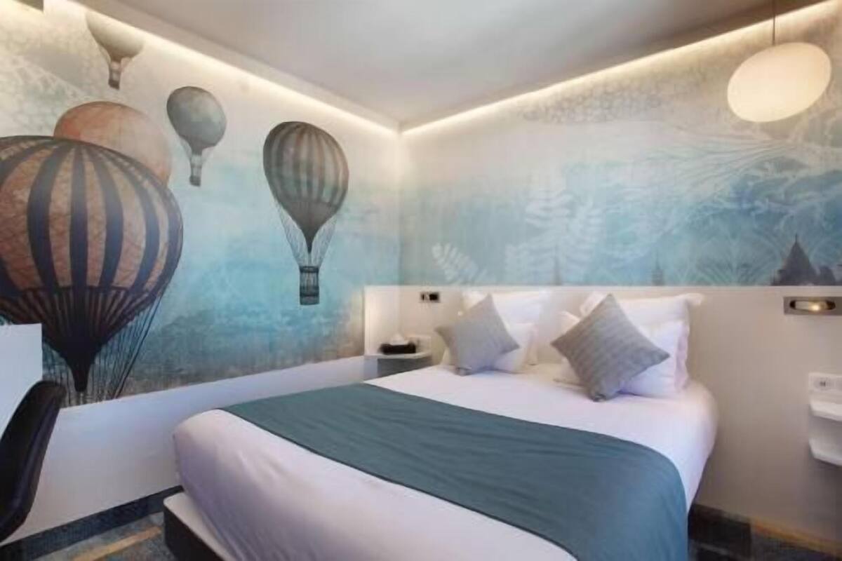 A hotel room with a bed, a nightstand, a chair, and a wall with a mural of hot air balloons.