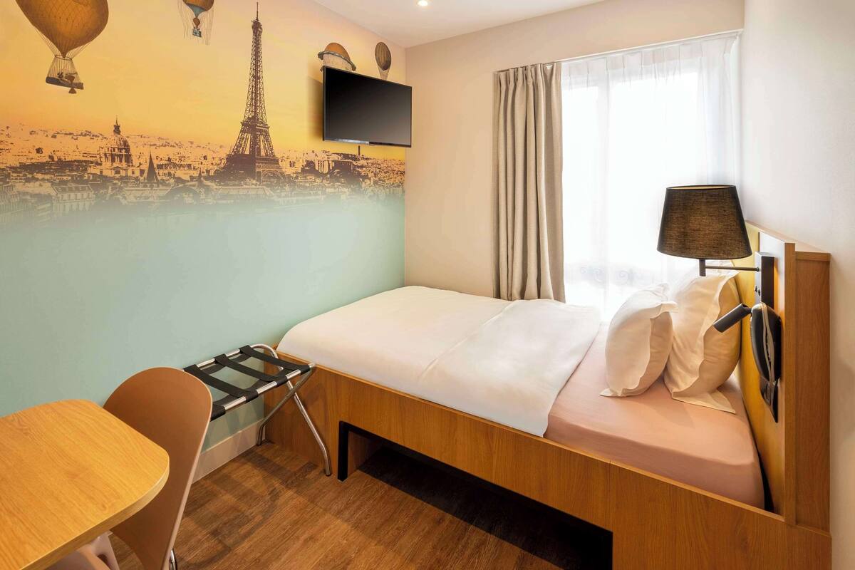 A hotel room with a bed, a desk, a chair, a TV mounted on the wall, and a mural of the Eiffel Tower.