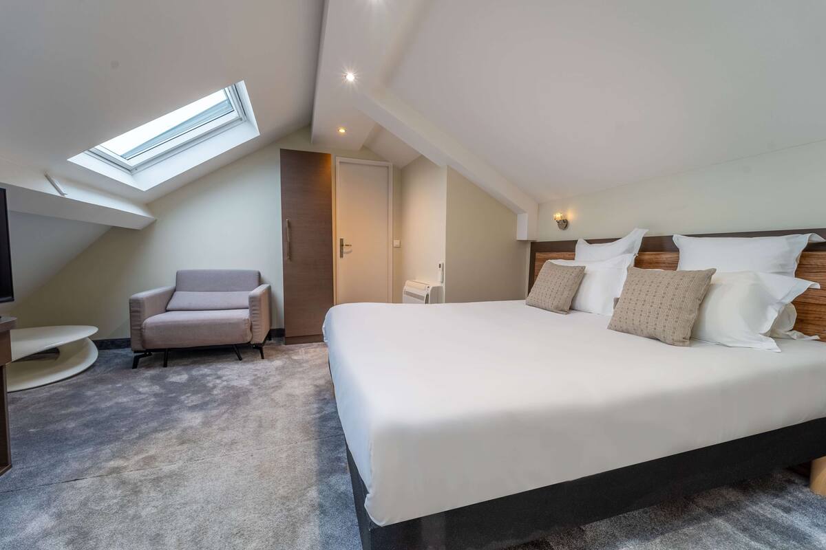 A bedroom with a skylight, a bed with multiple pillows, a chair, and a small table.