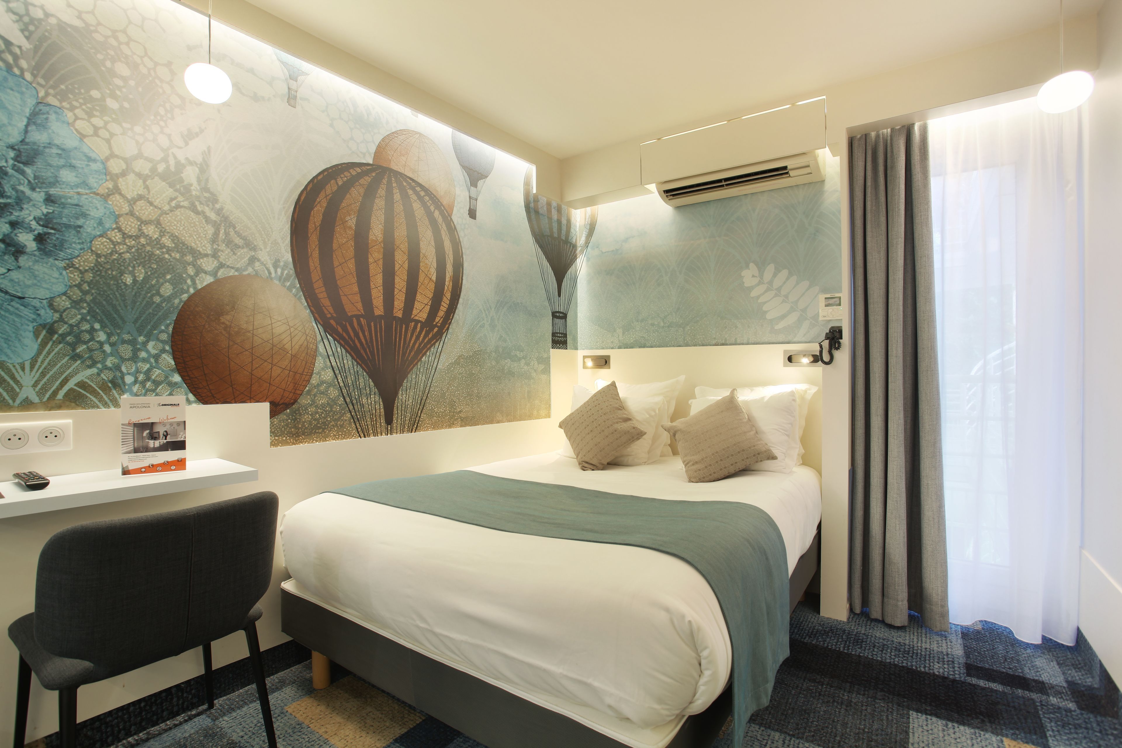 A hotel room with a bed, a desk, and a chair. There is a wall mural with hot air balloons.