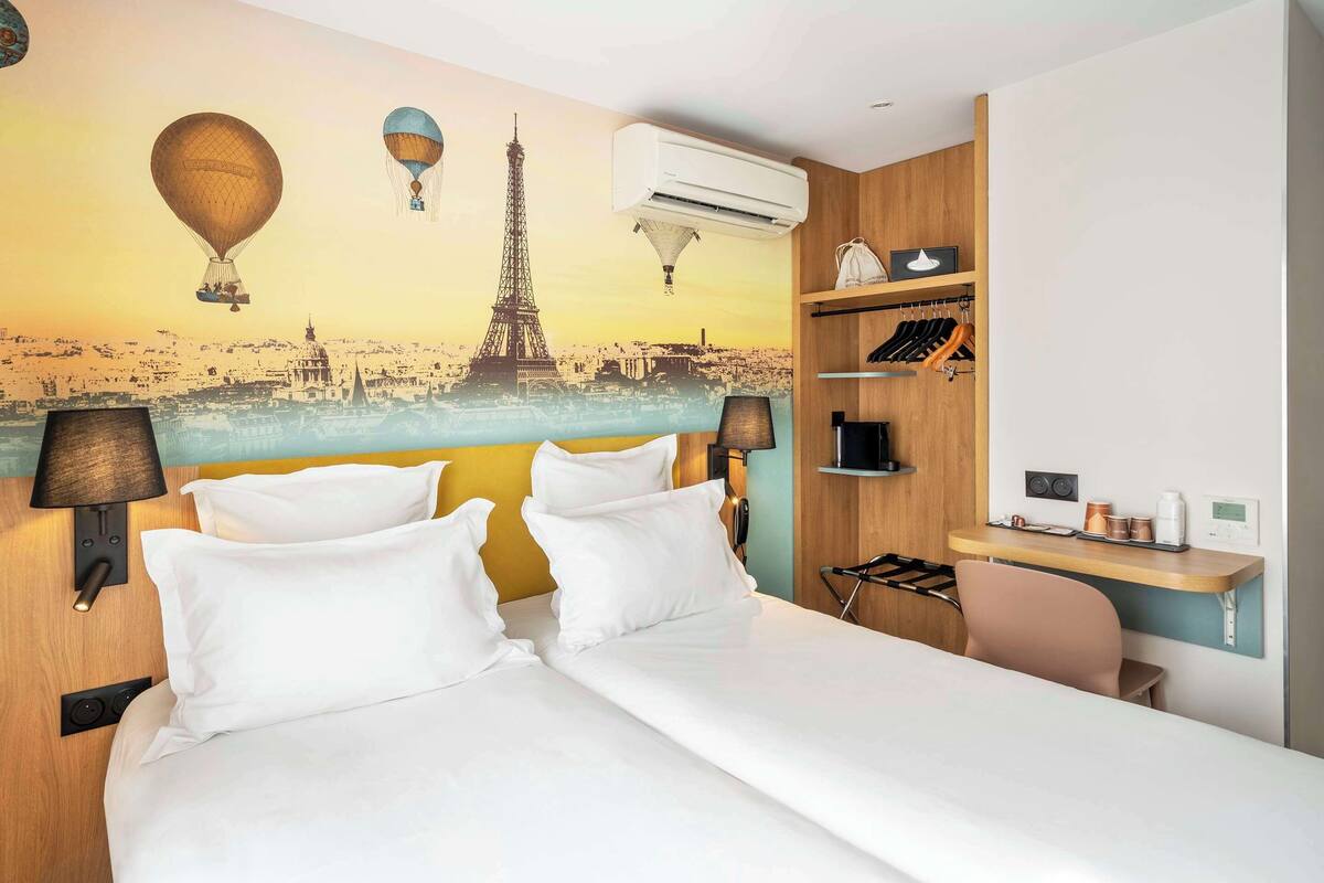 A hotel room with a bed, two bedside lamps, a wall-mounted air conditioner, a shelf with hanging items, and a chair.