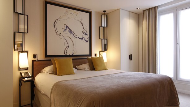 A hotel room with a large bed, two bedside lamps, a framed artwork above the bed, and a window with curtains. - Balmoral Champs Elysées (Paris)