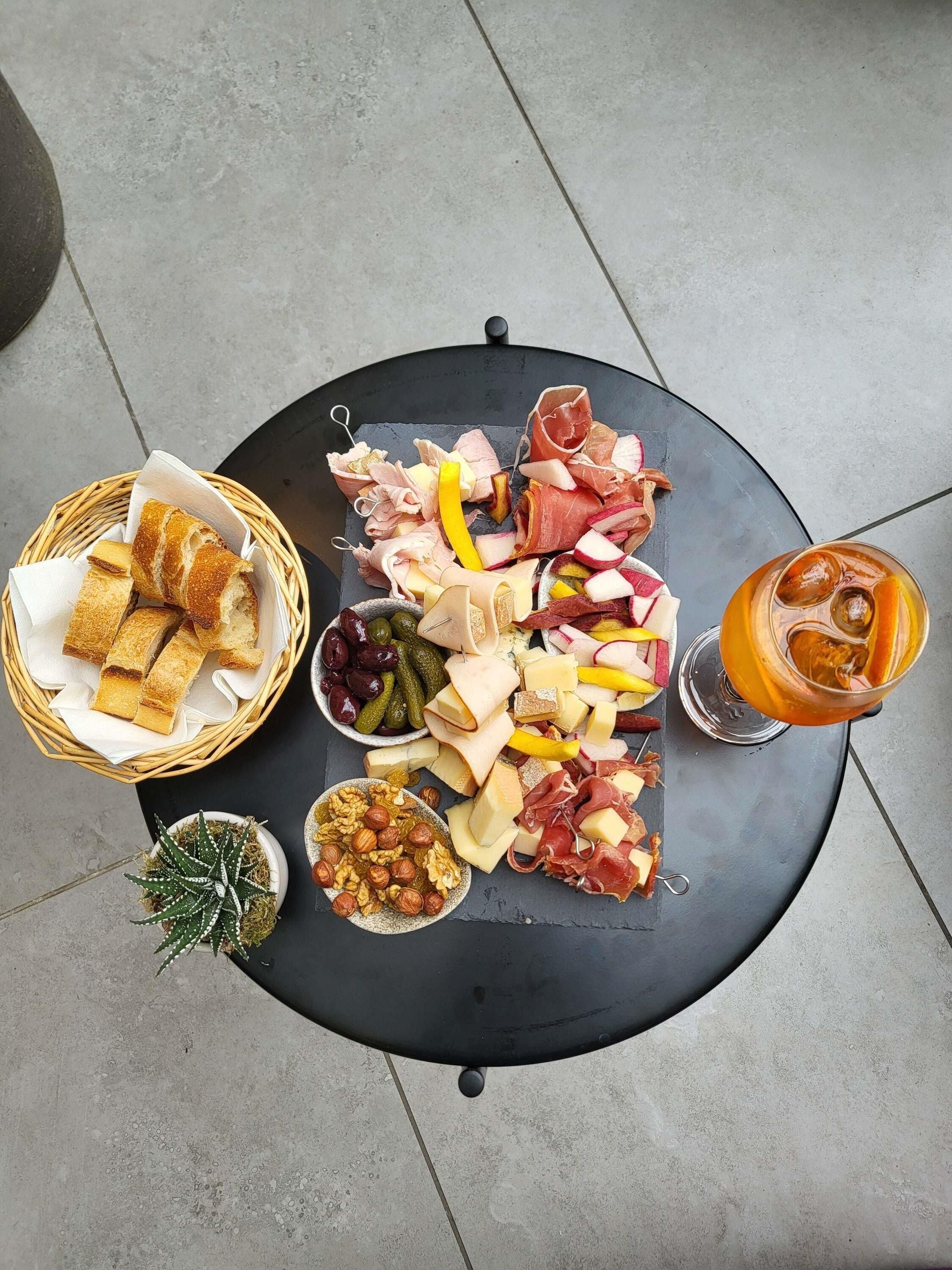 Una tabla de quesos y embutidos con diversos tipos de carne, queso y frutos secos, acompañada de un vaso de té helado, sobre una mesa redonda.