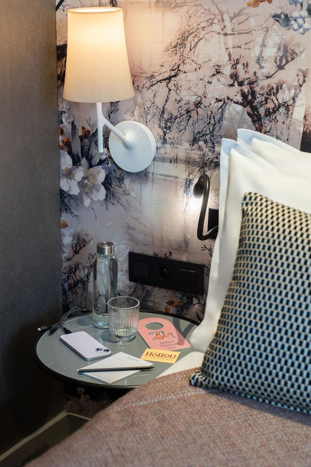 A hotel room with a bed, a bedside table with a lamp, a bottle, a glass, a notepad, and a card.
