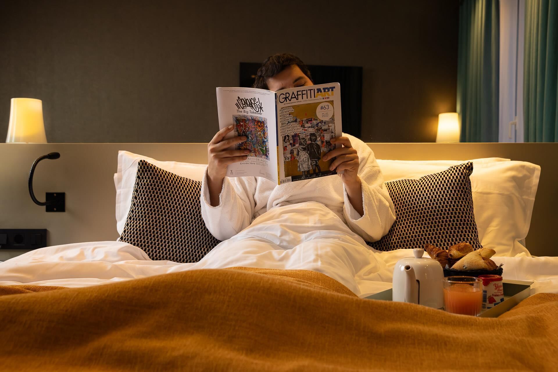 Eine Person liest ein Buch in einem Hotelzimmer. Es gibt ein Bett, Kissen, einen Nachttisch mit einem Wasserkocher und ein Tablett mit Croissants und Saft.