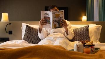 Una persona leyendo un libro en una habitación de hotel con cama, almohadas, mesita de noche con una tetera y una bandeja con cruasanes y jugo.