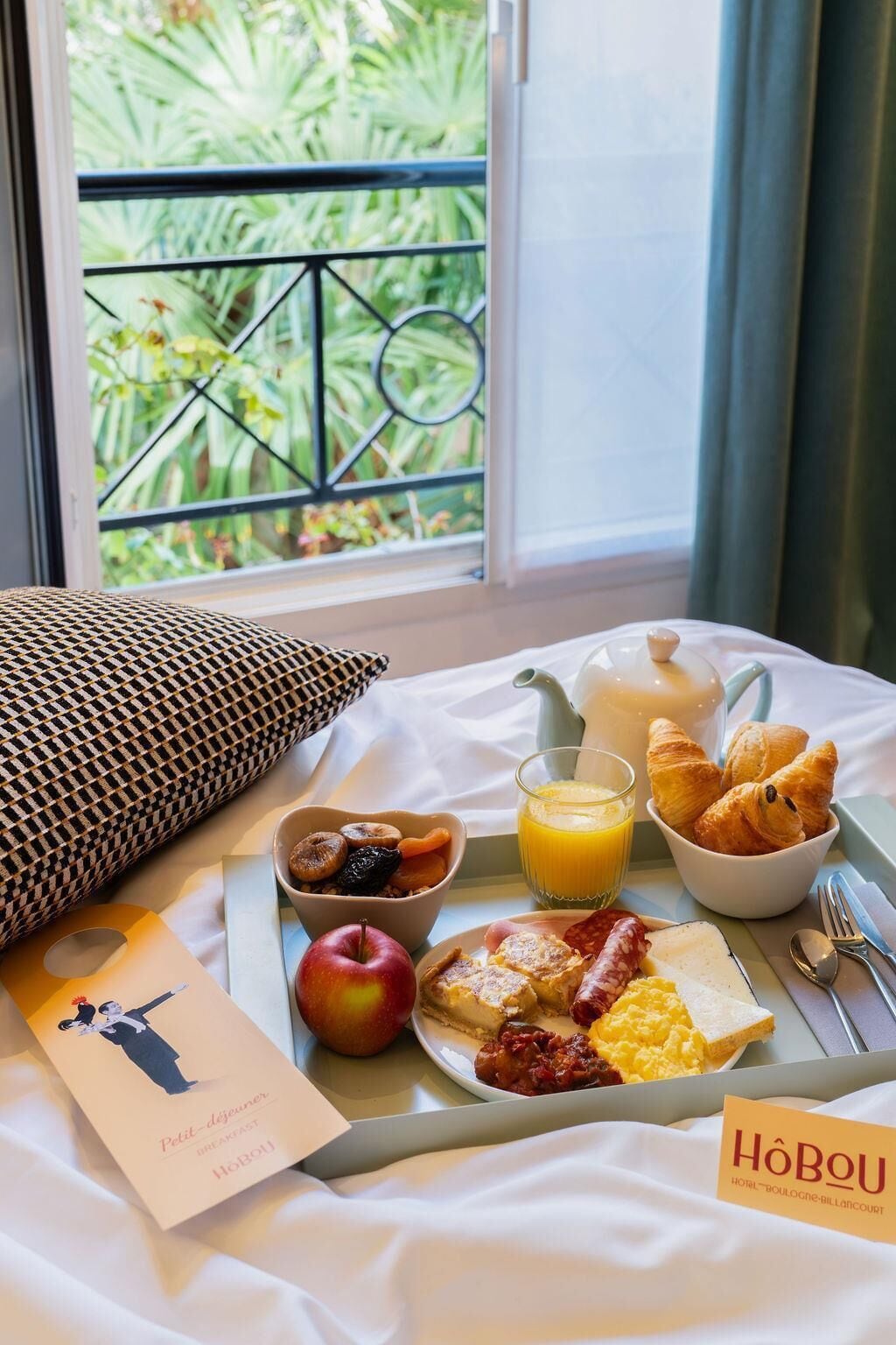 Bandeja de desayuno con cruasanes, fruta y huevos revueltos sobre una cama con vista a la vegetación.