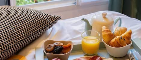 Bandeja de café da manhã com croissants, frutas e ovos mexidos sobre a cama, com vista para o verde lá fora.