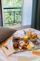 Bandeja de desayuno con cruasanes, fruta y huevos revueltos sobre una cama con vista a la vegetación.