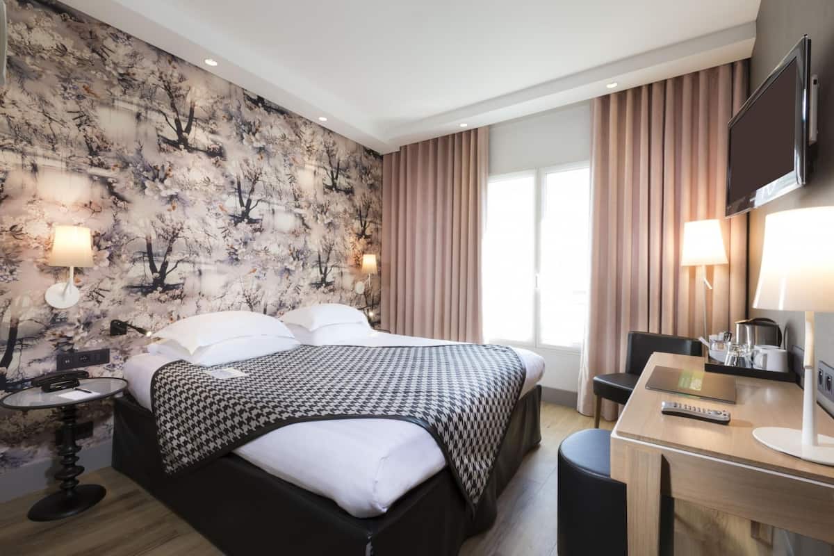 A hotel room with a bed, bedside table, lamp, desk with a chair, and a TV.