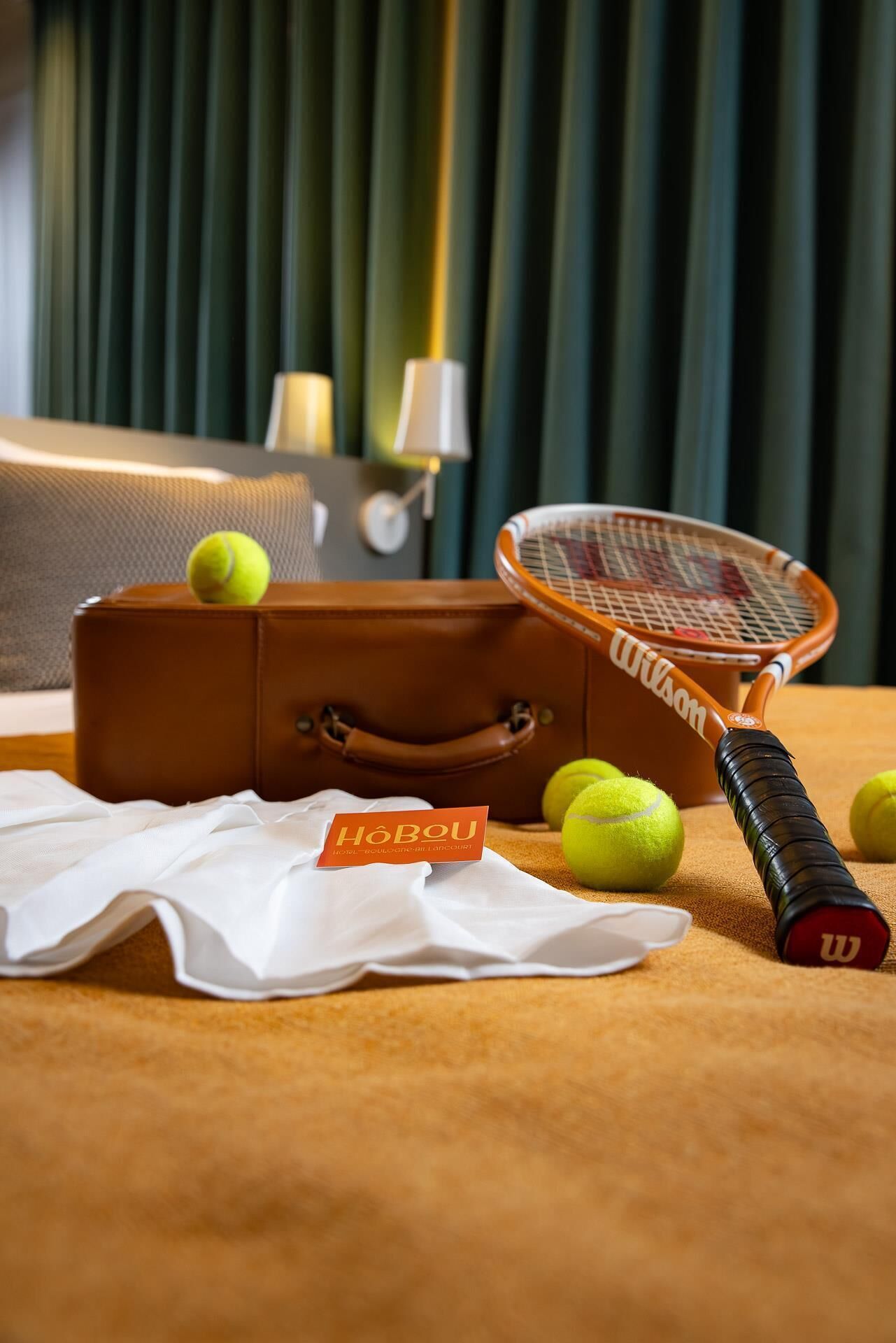 Une raquette de tennis, une valise et des balles de tennis sur un lit, avec une enseigne d’hôtel en arrière-plan.