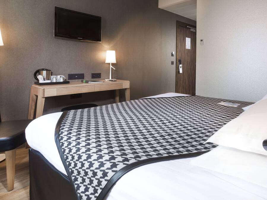 Quarto de hotel com cama, escrivaninha, cadeira, televisão e abajur.