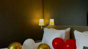 A hotel room with a bed, white pillows, and a bedside table with a lamp. There are balloons and rose petals on the bed.