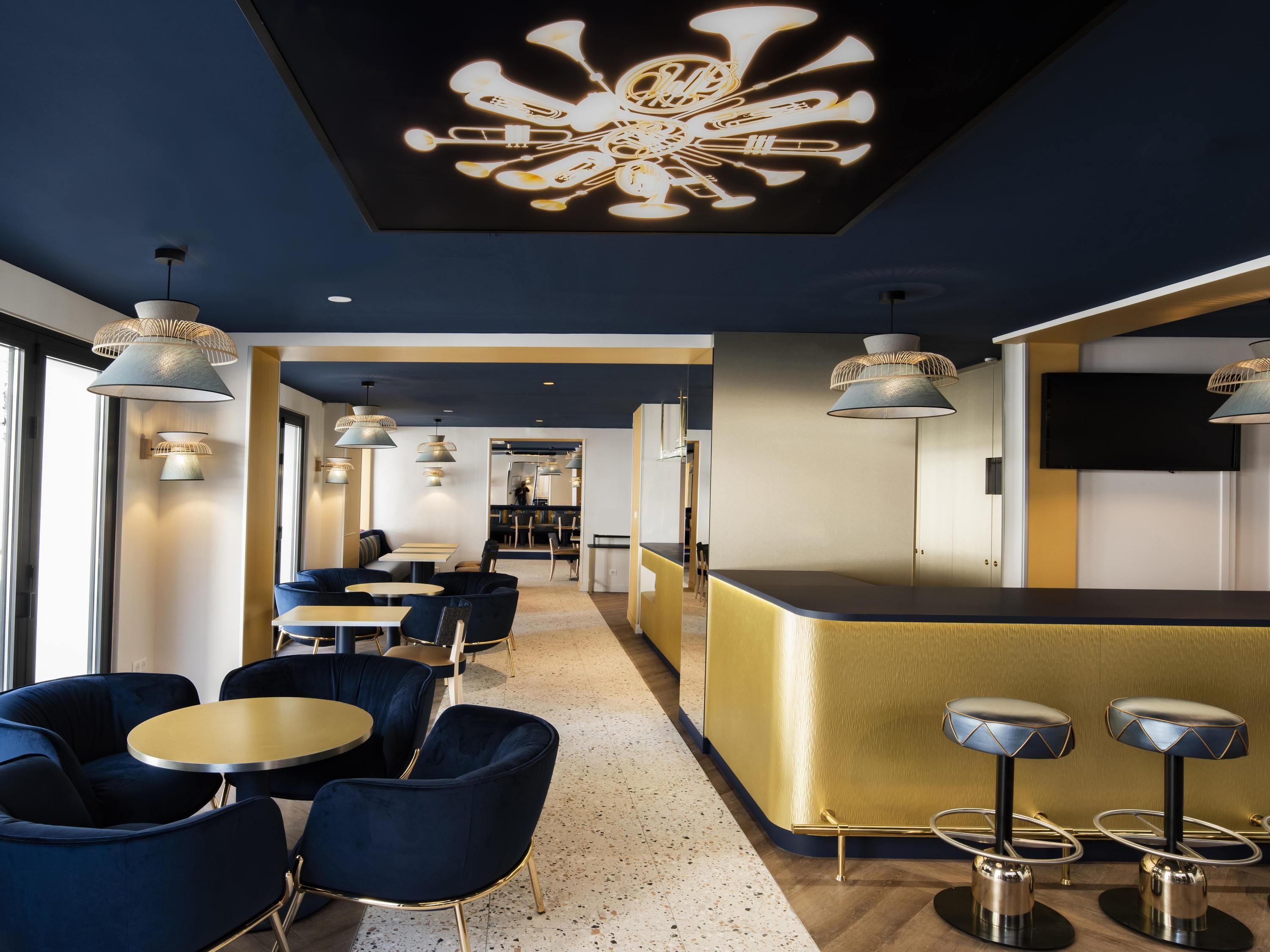 Een moderne hotellobby met ronde tafels, blauwe stoelen, een gouden receptiebalie en hanglampen.