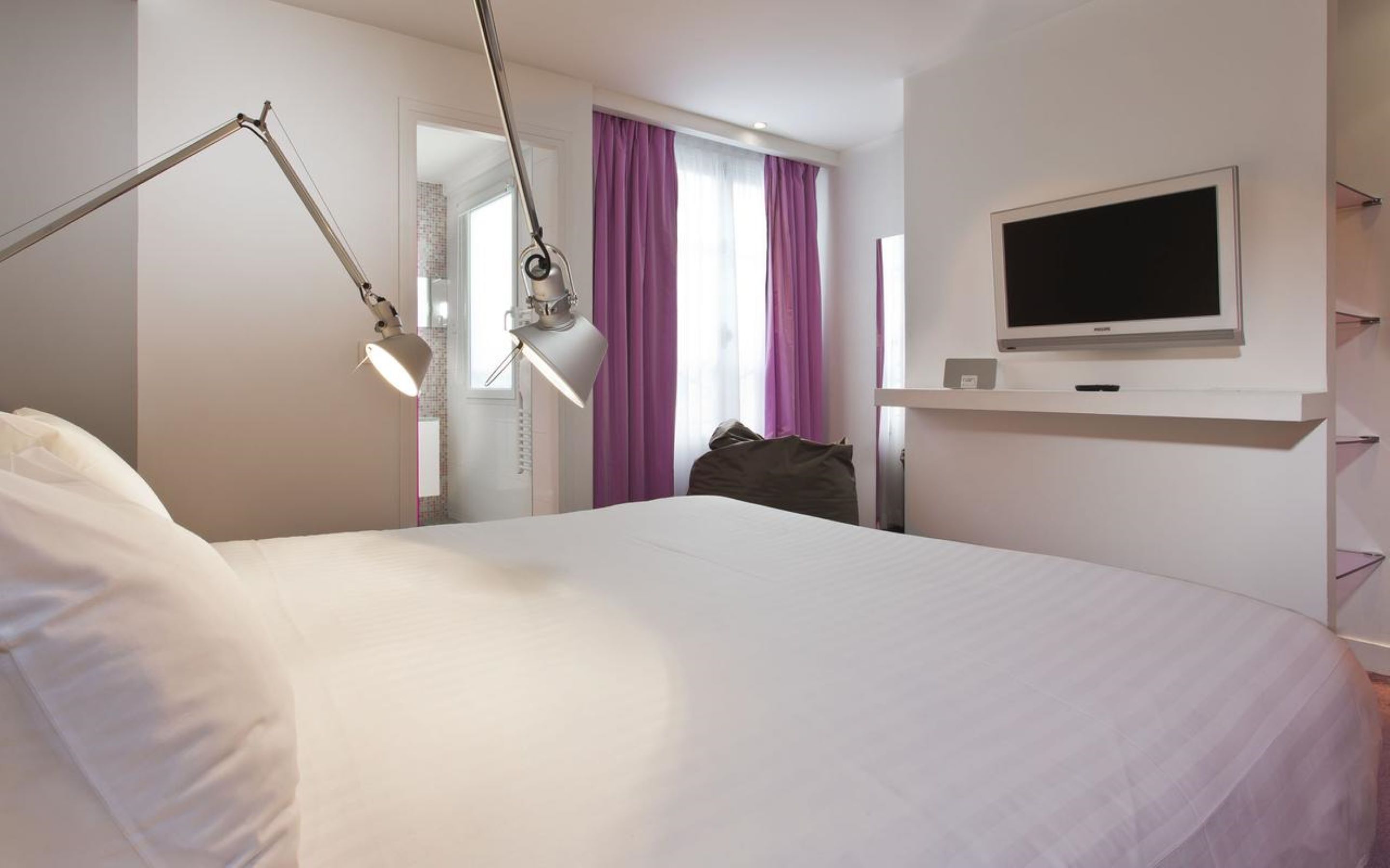 A hotel room with a bed, a desk, a TV, and a desk lamp.