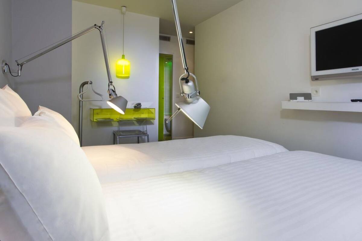 Habitación de hotel con una cama grande, un escritorio con silla, una lámpara de escritorio, un televisor montado en la pared y un estante pequeño.