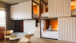 Ein modernes Hotelzimmer mit Stockbetten, einer kleinen Sitzecke mit Stühlen und einem Tisch sowie Blick auf die Stadt durch große Fenster.