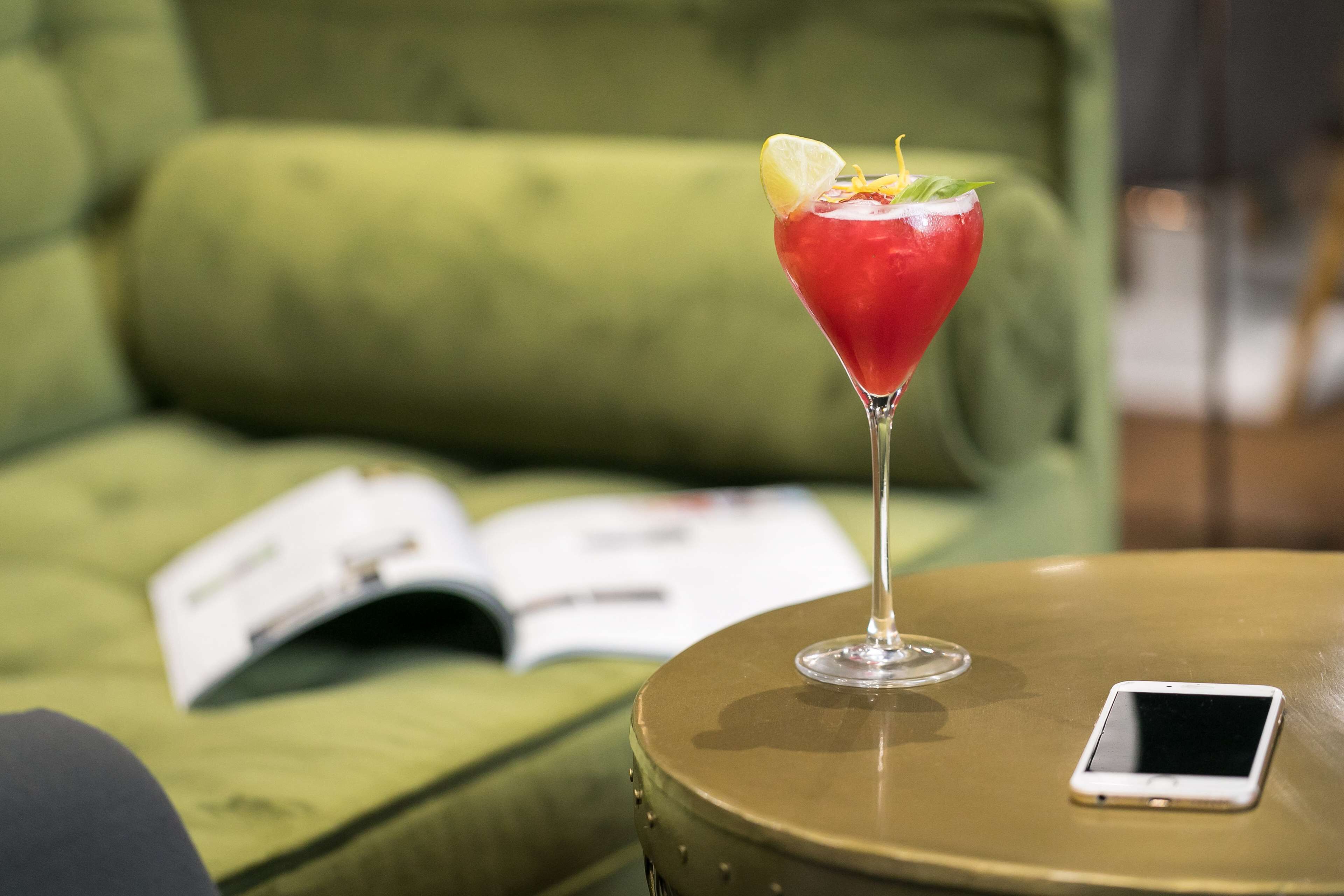 A martini glass with a garnish on a round table, a smartphone, and an open book on a green sofa.