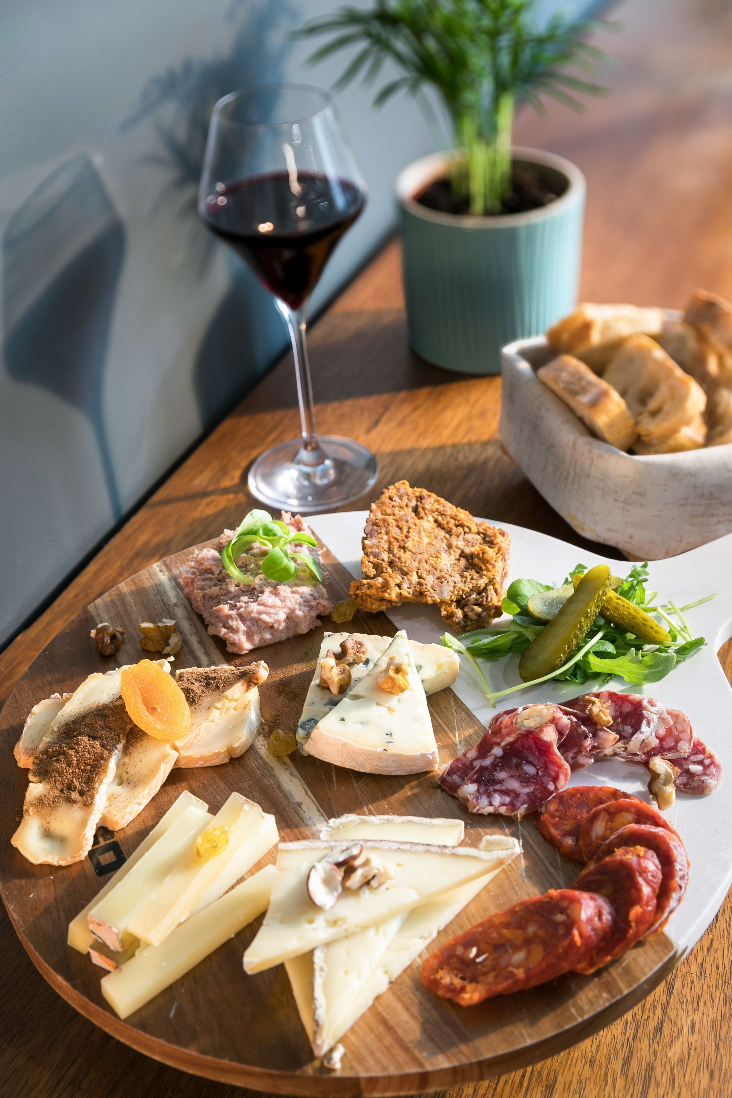 A charcuterie board with various cheeses, meats, and bread, accompanied by a glass of red wine and a potted plant.