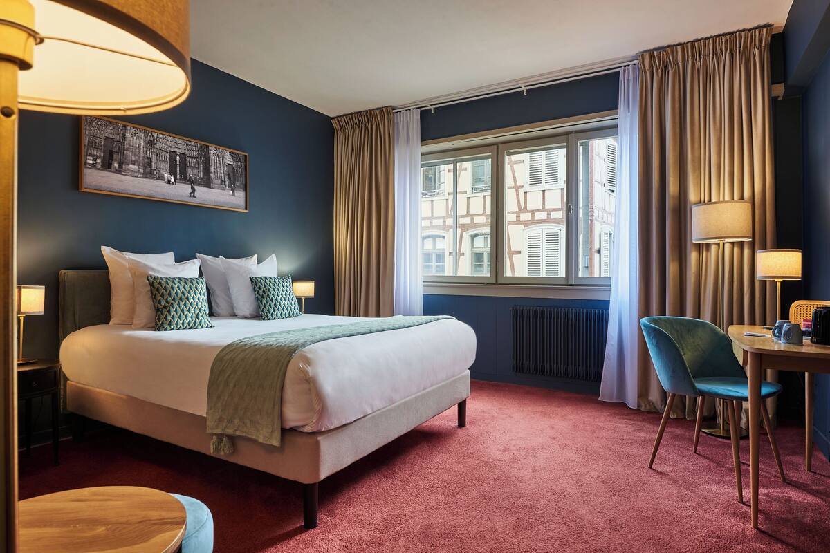 Ein Hotelzimmer mit einem großen Bett, einem Schreibtisch, einem Sessel, einem Fenster mit Vorhängen und einer Lampe.