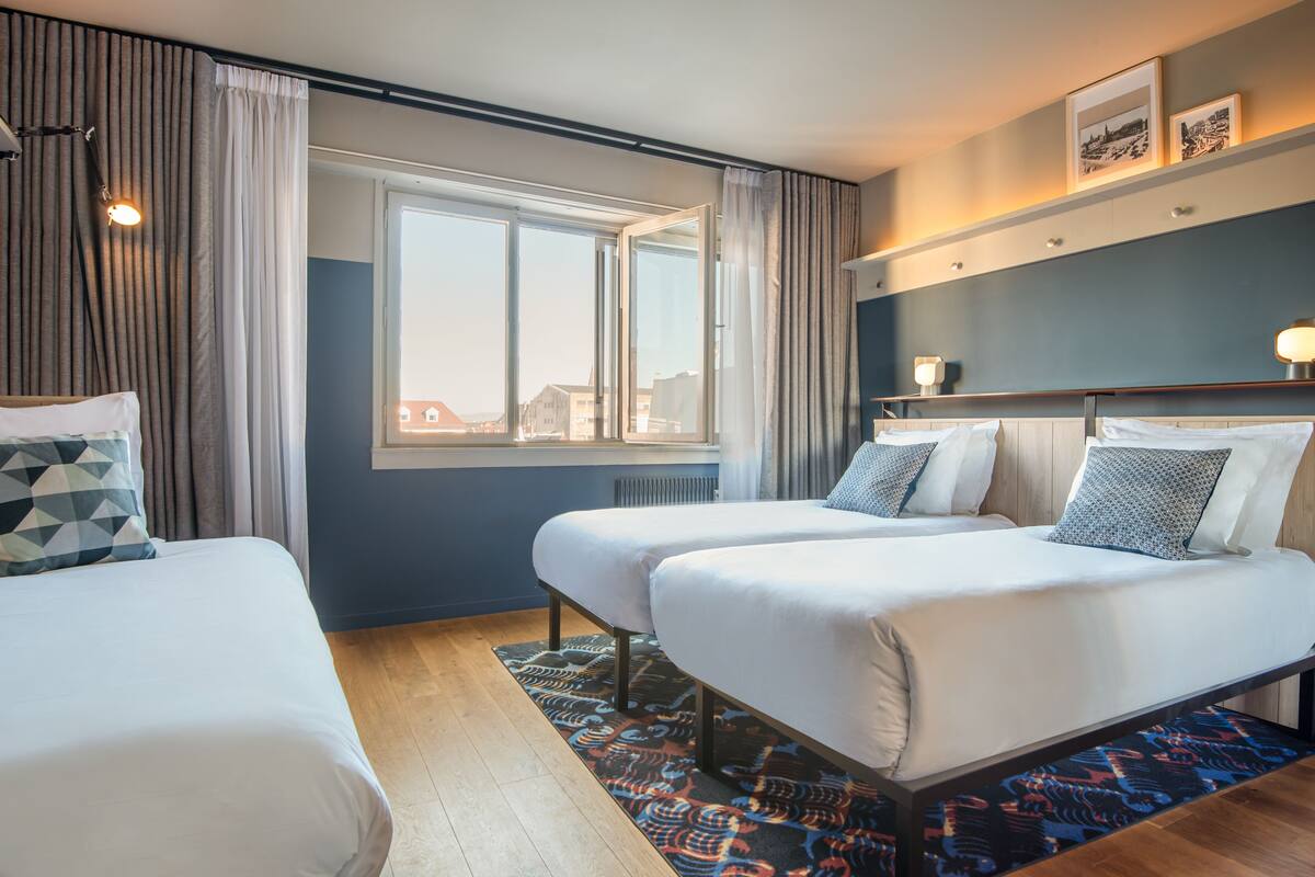 Ein Hotelzimmer mit zwei Betten, einem großen Fenster, Blick auf Gebäude und einem gemusterten Teppich.