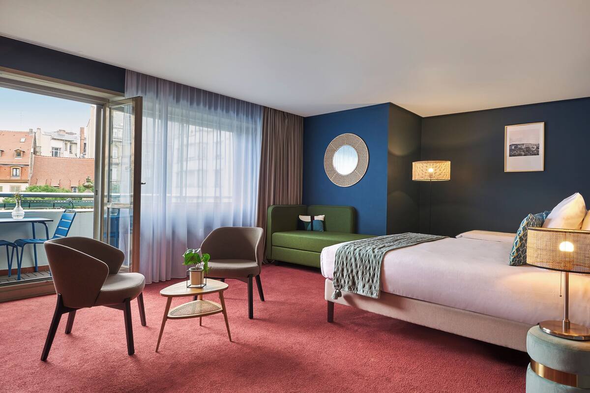 Ein Hotelzimmer mit einem Bett, zwei Stühlen, einem kleinen Tisch mit einer Pflanze, einem runden Spiegel an der Wand und Blick auf Gebäude durch das Fenster.