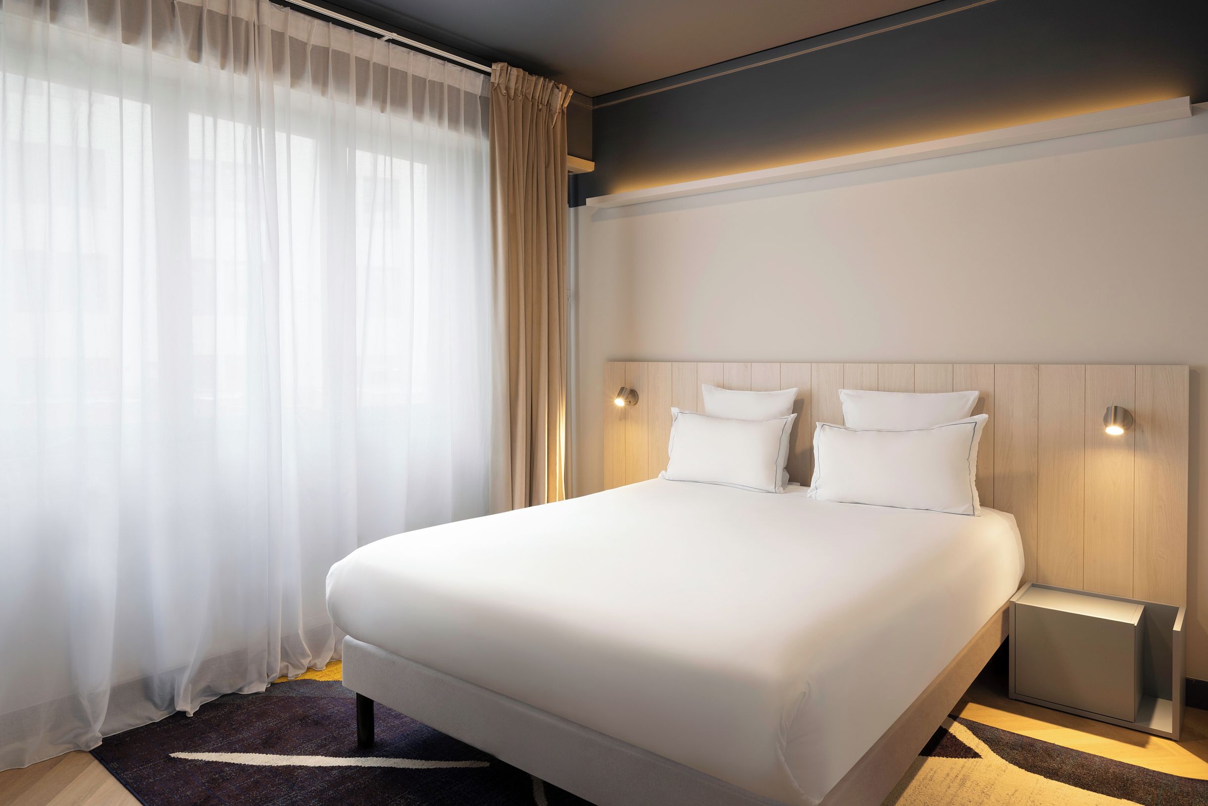 A hotel room with a large bed, two bedside tables, and a window with sheer curtains.