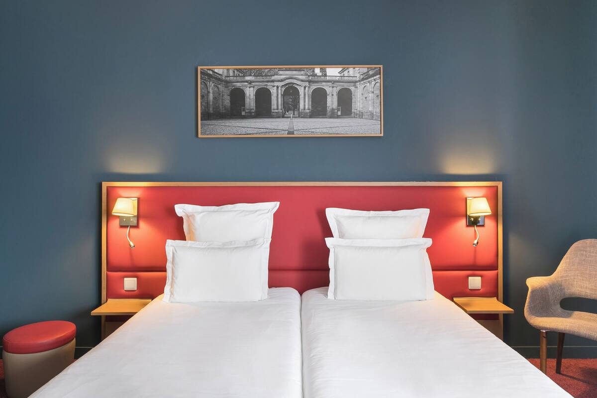 Ein Hotelzimmer mit rotem Kopfteil, zwei Betten mit weißen Laken, einer roten Mülleimer und einem gerahmten Bild an der Wand.