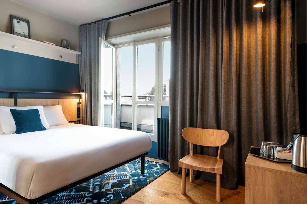 Ein Hotelzimmer mit einem großen Bett, einem Holzstuhl, einem Schreibtisch mit Wasserkocher und Blick auf Gebäude durch die Fenster.