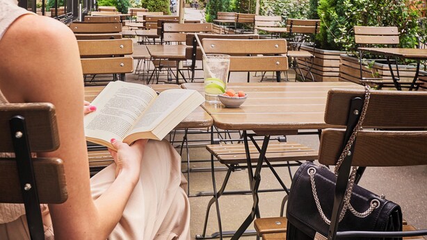 Eine Frau liest ein Buch in einem Straßencafé mit Holztischen und beigefarbenen Stühlen, unter großen Sonnenschirmen, mit Gebäuden im Hintergrund.