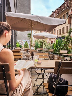 Een vrouw leest een boek in een openlucht café met houten tafels en beige stoelen, onder grote parasols, met gebouwen op de achtergrond.