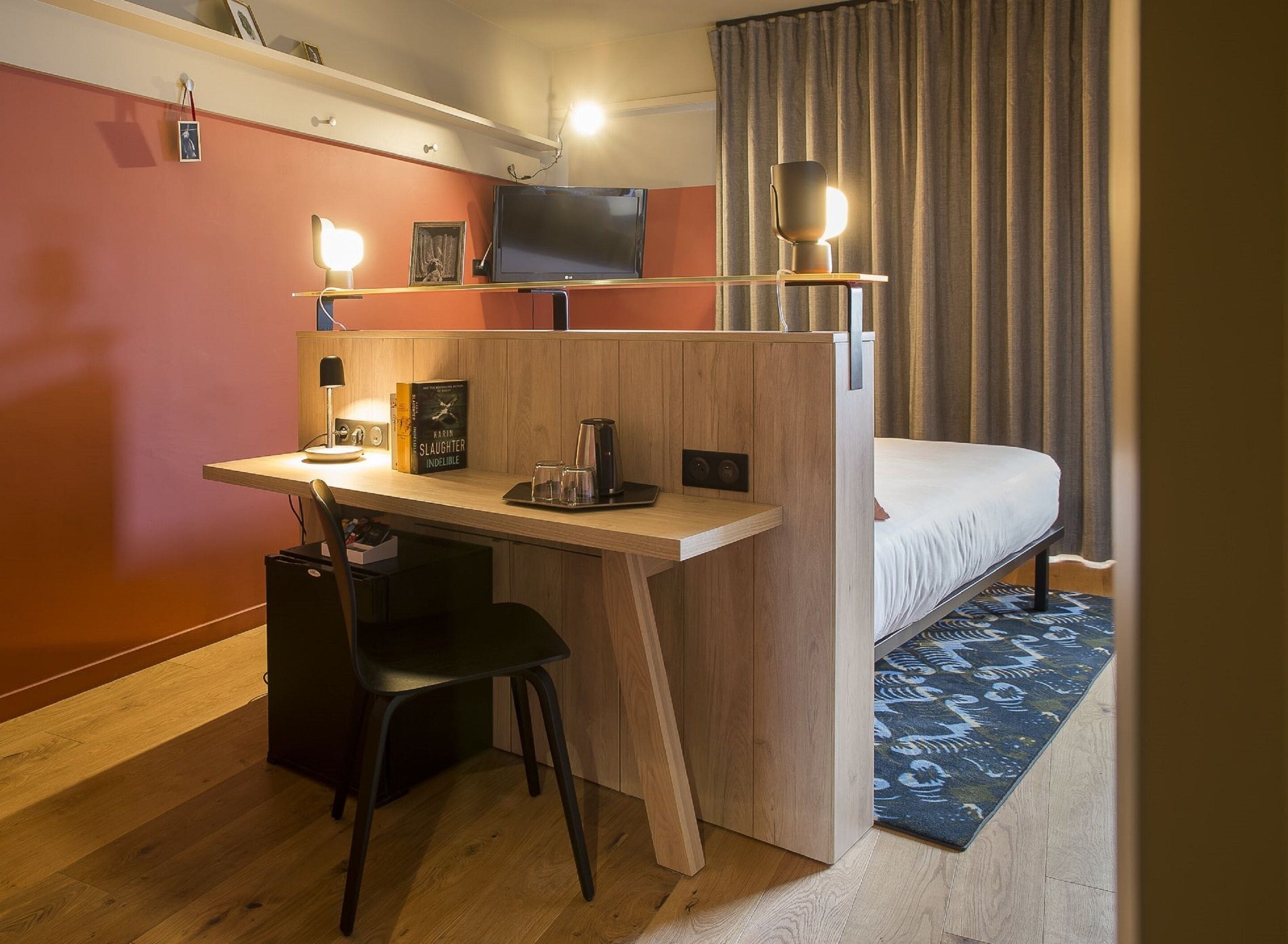 A compact hotel room with a wooden desk, a single bed, a small TV, and a desk lamp.