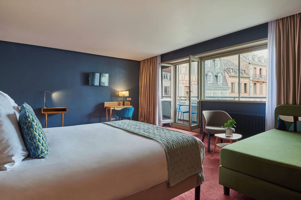 Ein Hotelzimmer mit einem großen Bett, einem Schreibtisch mit Stuhl, einem kleinen Tisch mit Pflanze und Blick auf Gebäude durch ein großes Fenster.