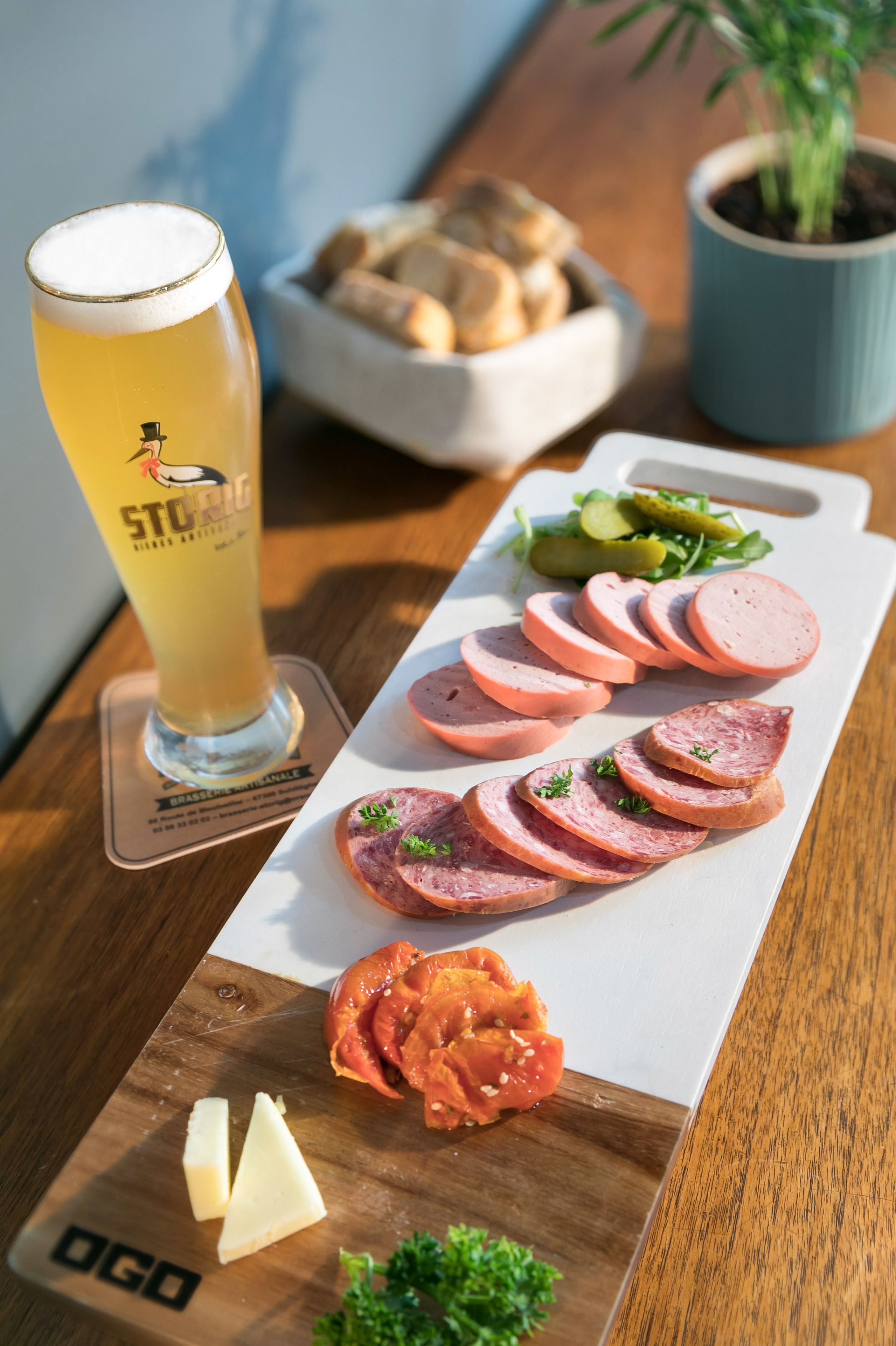 Una mesa de madera con una cerveza, salchichas en rodajas, pepinillos, queso y una planta en maceta.