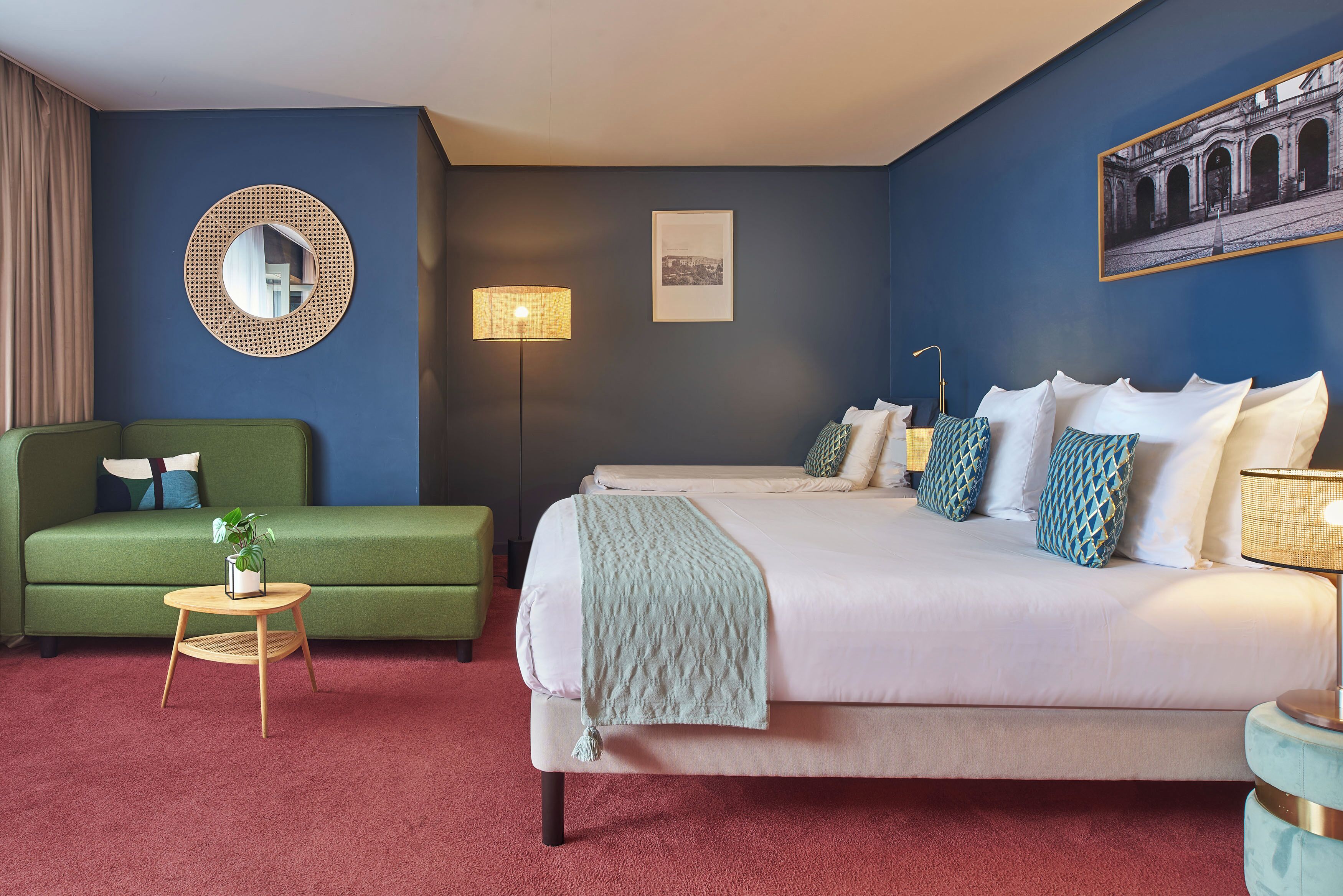 A hotel room with a bed, a green sofa, a small round table with a plant, a round mirror, and a wall-mounted lamp.