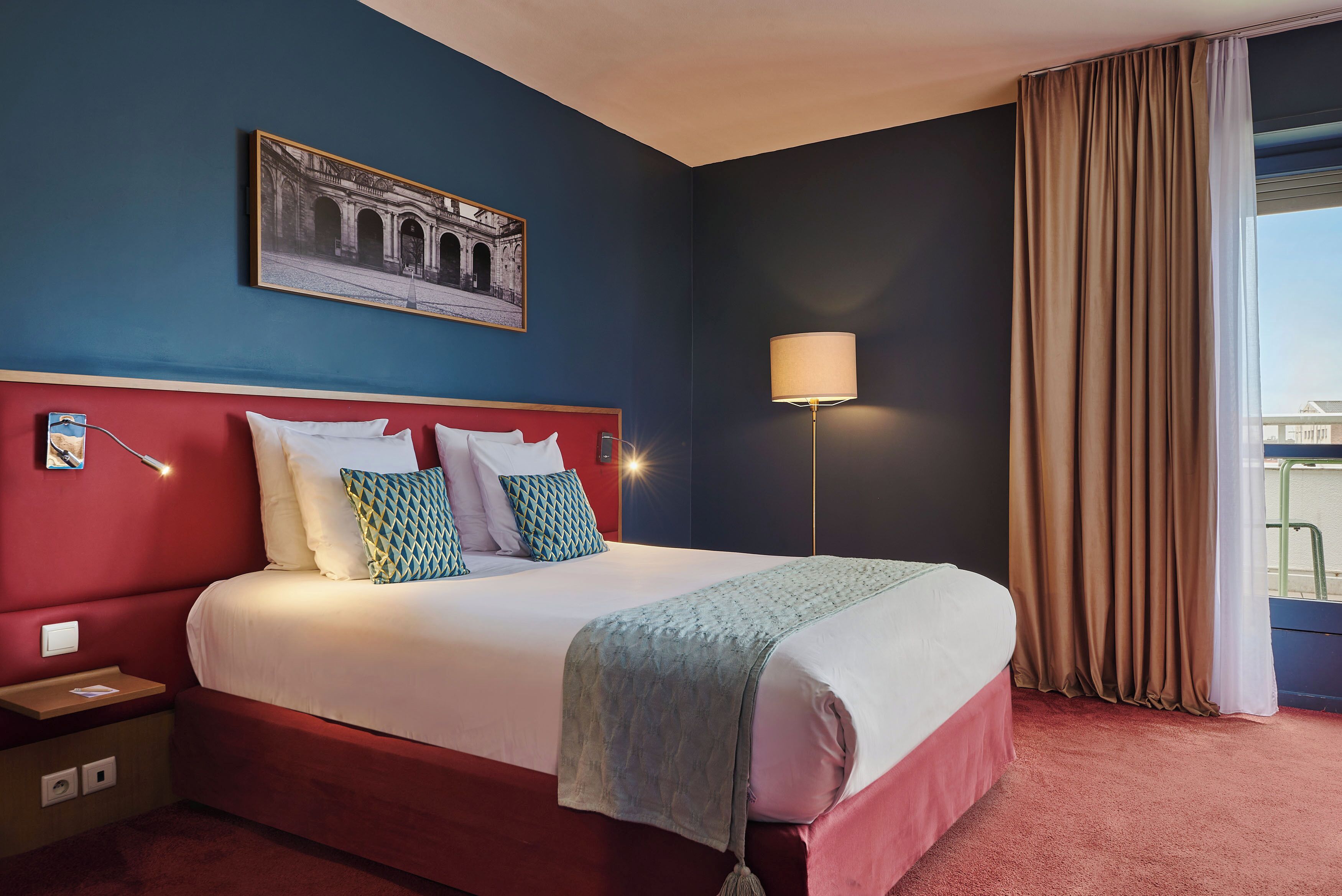 A hotel room with a large bed, a nightstand, a lamp, a window with curtains, and a framed picture on the wall.