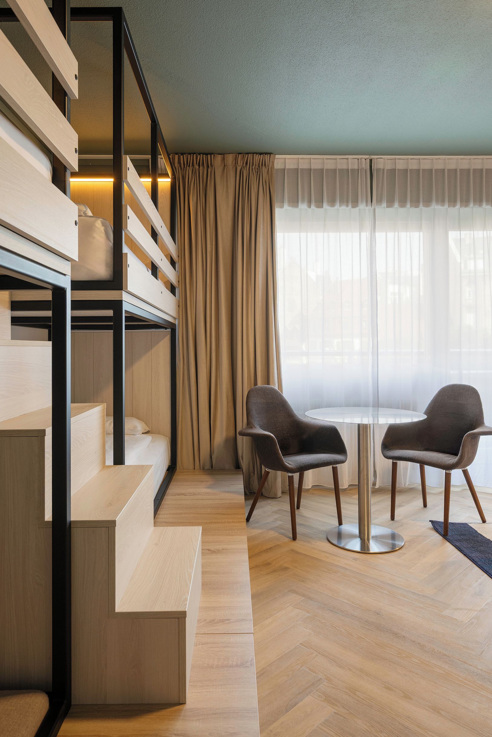 A modern hotel room with a wooden staircase, a bed, two armchairs, a round table, and a large window with sheer curtains.